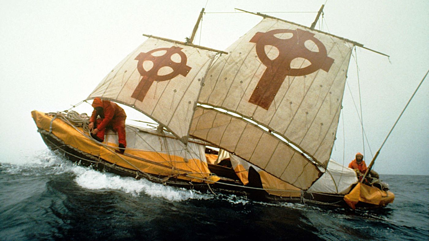 A leather-hulled currach under full sail in heavy Atlantic seas, its sails marked with Celtic crosses and two crew in orange oilskins