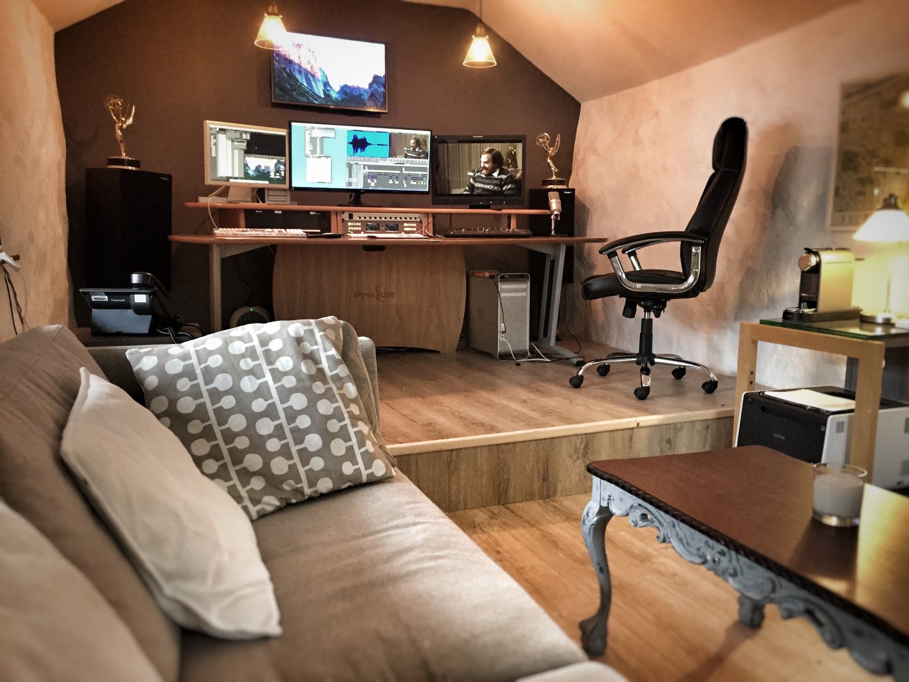 Bob Caldwell's edit suite: three monitors on a curved wooden desk under pendant lights, with a sofa in the foreground